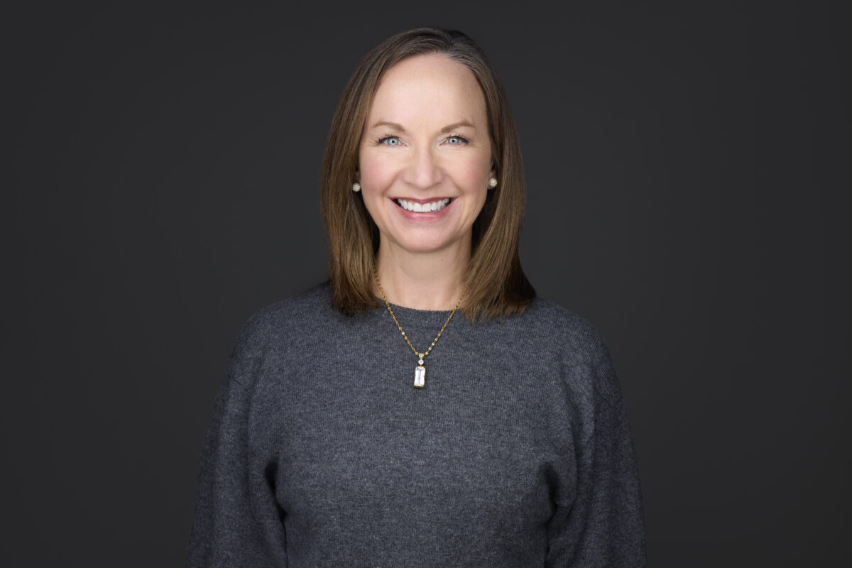 Woman with medium lengthe brown hair and wearing a grey sweater, facing the camera and smiling. 